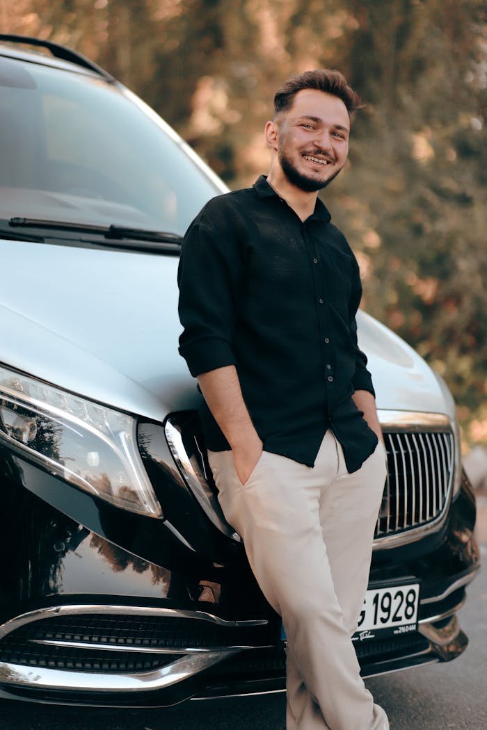 Casually dressed man smiling and leaning against a luxury car on a sunny day.